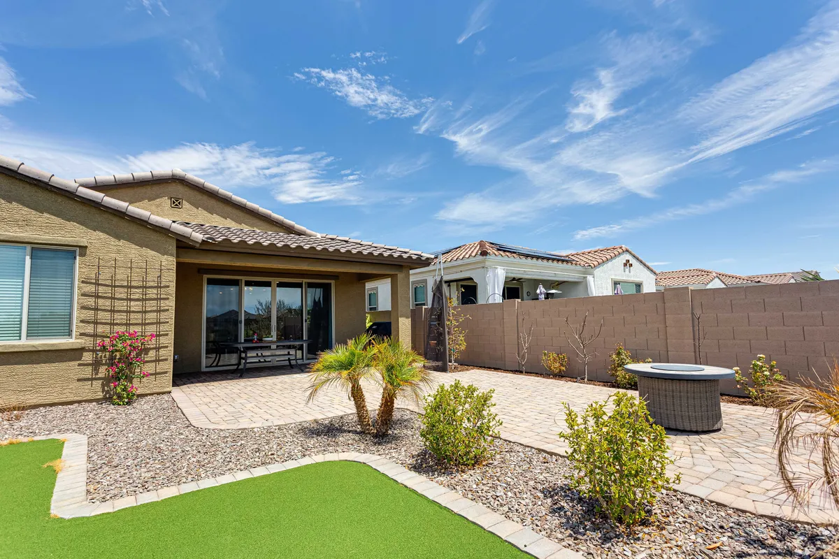 A tranquil backyard patio area with comfortable seating around a fire pit, surrounded by lush greenery and decorative stones. The area is framed by vibrant landscaping and hints at beautiful sunset views.