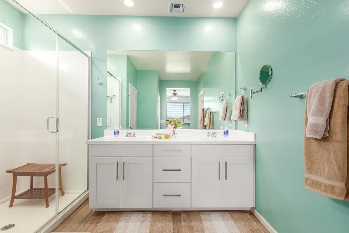 A spacious, spa-inspired bathroom featuring a double sink vanity, a glass shower enclosure, a wooden stool, and a large mirror. The layout is bright and serene, evoking a luxurious, calming experience ideal for restorative routines.