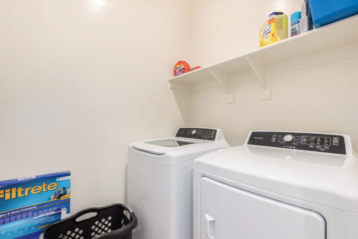 A bright, practical laundry room featuring a modern washing machine and drying machine, with a shelf above stocked with laundry supplies. The clean and organized setup conveys everyday function and convenience.