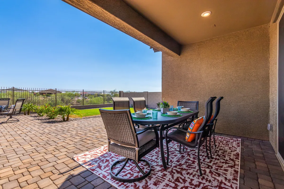 An exterior patio scene featuring a stylish dining table and chairs set on a decorative rug, with a sweeping landscape view beyond. The setup suggests an ideal spot for outdoor dining, socializing, and relaxing in the Arizona sunshine.