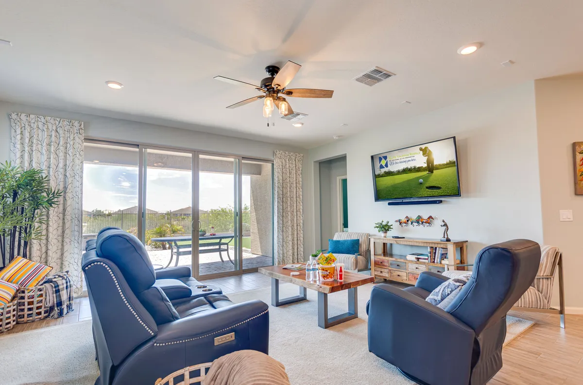The image focuses on a comfortable living area with blue recliners, a wooden coffee table, and large windows revealing outdoor views. A large TV is featured, reinforcing the entertainment aspect. The casual, welcoming setup is perfect for family relaxation, social gatherings, or quiet mornings.