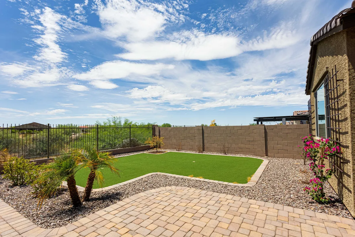A peaceful backyard featuring a green lawn, vibrant flowering plants, a paved pathway, and a wooden fence, all bathed in natural sunlight. The space feels inviting for both relaxation and outdoor recreation.